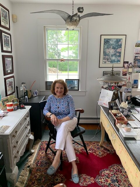 Darby Hobbs sitting in her cozy office with a desk, a colorful rug, and a wooden goose "flying" above her.